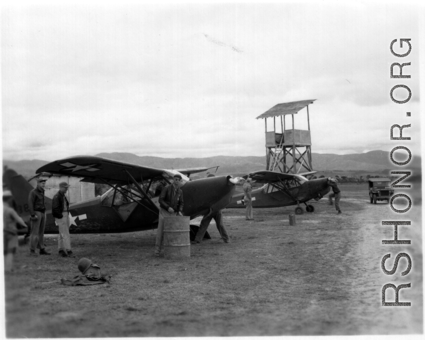 A pair of Stinson L-5's at a base in China, possibly Yangkai.  Notice the man in the background starting the plane, and the simplified control tower.