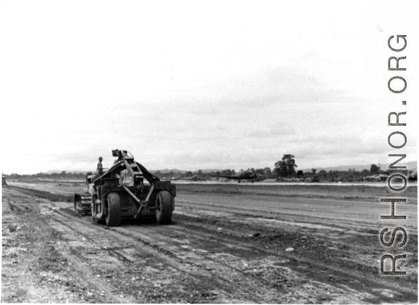 Mechanical grader flattens out a runway in the CBI during WWII, while C-47 moves in the background.