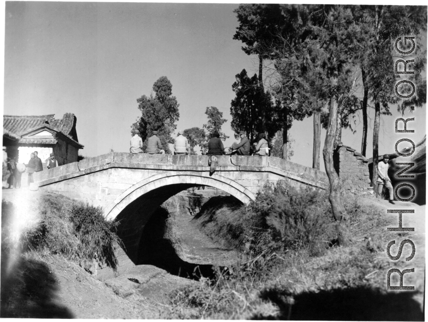 Chinese civilians and Nationalist soldiers pass the time on a small bridge in Yunnan, China, probably near Kunming or Yangkai base. During WWII.  From the collection of Hal Geer.