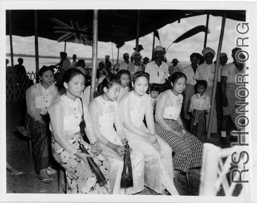 Local people in Burma near the 797th Engineer Forestry Company--Ladies in their finery sit under a covering, with a Union Jack flying in the background, in Burma.  During WWII.