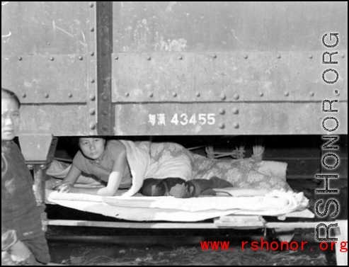 A Chinese refugee has made a tenuous and dangerous bed on the framework hanging below at train car. At the train station in Liuzhou during WWII, in the fall of 1944, as the Japanese advanced during the Ichigo campaign.