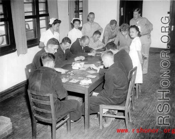 A crowd of servicemen in robes and in uniforms sit around a table playing bingo, with three Chinese nurses looking on in curiosity.