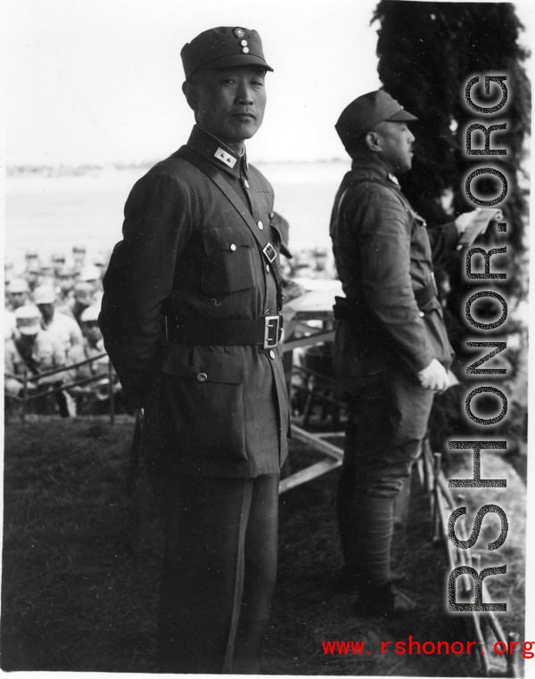 Chinese Lt. General Du Yuming, commander of Nationalist 5th Corps (第五集团军总司令兼昆明防守司令杜聿明) looks at American photographer, with Gen. Wei Lihuang (卫立煌) facing assembled troops, and another person holding a sheet of paper, on stage during rally.