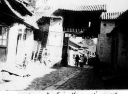 Village street in Yunnanyi, China, during WWII, with gate having a sign for Shanghai Man Hairstyling (上海男美发室), and the Almond Flower Tea Room (杏花茶室）in the shadow on the right. 1944.