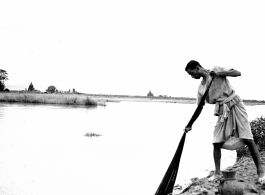 A man fishes with a next from the bank of a river. Probably in India. During WWII.