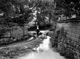 A man in rural Yunnan province, China, carries a heavy weight on a should pole while as western woman (in light colored dress and hat) looks on. During WWII.