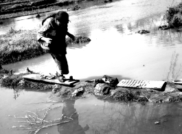 Local people in China: Elderly woman with bound feet crosses a precarious pathway of mud, boards, and stones. During WWII.  From the collection of Eugene T. Wozniak.
