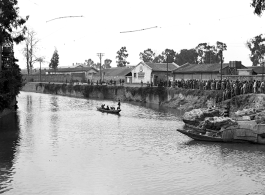 A boat set up to fish using cormorants in a canal in Kunming, while a large group of people have gathered, apparently watching the cormorant fishermen.   In Yunnan province, China, during WW