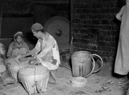 A family kneeling and working at a grinding mill in China during WWII, while a customer stands waiting. Note the large grinding wheel in motion behind them.