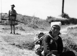 An elderly Chinese woman takes her lunch somewhere outside of Kunming, while a Nationalist guard is in the background. During WWII.