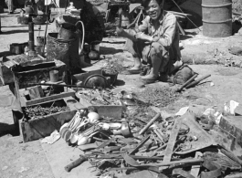 An ironmonger with his wares (including spoons made with the exotic metal aluminum, scavenged from crashed airplanes). Likely in Yunnan province, China, most likely around the Luliang air base area.