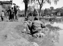 Tall American photographer lugs camera and tripod through village in Yunnan, China, during WWII, while people go about daily life.