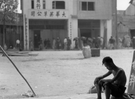 A tired looking young man sits in the opening to a stall advertising car parts, in Kunming, China, during WWII.
