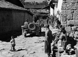 GI drives jeep in village lane among adobe brick houses in Yunnan, during WWII.