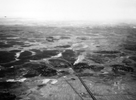 A B-25 flies above a smoking village on a railway in SW China, French Indochina, or Burma, in the CBI, during WWII.