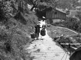 A man carries large packages up a steep mountain path using a shoulder pole in China during WWII.