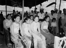 Local people in Burma near the 797th Engineer Forestry Company--Ladies in their finery sit under a covering, with a Union Jack flying in the background, in Burma.  During WWII.