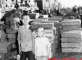 Two refugee children pose for the photographer before crates of supplies. At the train station in Liuzhou during WWII, in the fall of 1944, as the Japanese advanced during the Ichigo campaign.