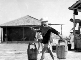 A Chinese staff person at an American base in China during WWII carries buckets on a shoulder pole.