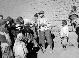 A photographer interacting in a village in Yunnan province.  He's a corporal, so he hasn't been in the service very long. The pith helmet he's wearing is something GIs frequently bought in India on their way into China. The Speed Graphic he's shooting is a late model (earlier Graphics did not did not have the Military ID plate on the bottom of the door).