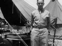 An American stands next to a tent with bed cots, in the CBI, during WWII.