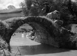 A rural woman shouldering baskets crosses a worn footbridge in China.  From the collection of Eugene T. Wozniak.