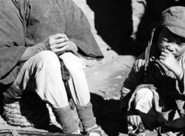 Local people in China: An elderly woman---possibly working as a petty seamstress doing minor repairs on shoes or cloth items---sits with child. During WWII.