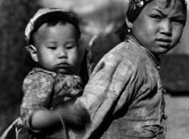 Local people in China: An older sister carries a younger child piggyback as part of her daily responsibilities. During WWII.