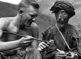 A GI inspects smoking pipe of local man in China, while local man holds western-style pipe. During WWII.  Photo by Syd Greenberg.