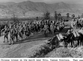 Chinese troops march near Mitu, Yunnan province, during WWII. They pass pack mules with wooden saddles as an American liaison officer and GIs (right center beyond mules) watch the passing parade.  US Signal Corps Photo.