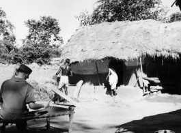 Soldier sitting on cot while local people go about chores.  Scenes in India witnessed by American GIs during WWII. For many Americans of that era, with their limited experience traveling, the everyday sights and sounds overseas were new, intriguing, and photo worthy.