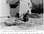 A GI points his carbine down the narrow space between two homes in a Chinese village during WWII, as a local village woman threshes grain.