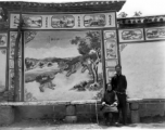 An elderly woman and man pose before a traditional decorative painted wall in SW China during WWII.