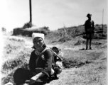 A woman in Yunnan province, China, rests to eat a meal, during the hard days of WWII.  Note the foot container on her lap and the chopsticks in her hands. A soldier with gun stands in the background.  From the collection of Eugene T. Wozniak.