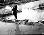 Local people in China: Elderly woman with bound feet crosses a precarious pathway of mud, boards, and stones. During WWII.  From the collection of Eugene T. Wozniak.