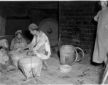 A family kneeling and working at a grinding mill in China during WWII, while a customer stands waiting. Note the large grinding wheel in motion behind them.