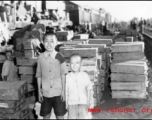 Two refugee children pose for the photographer before crates of supplies. At the train station in Liuzhou during WWII, in the fall of 1944, as the Japanese advanced during the Ichigo campaign.