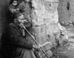 Local people in Yunnan province, China: Elderly man sits in village doorway and smokes from very long pipe. During WWII.