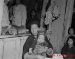 A woman holds a small child in a shop in Yunnan, China, during the Second World War.  Note the straw sandals hanging on the shop window in the back.