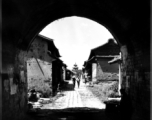 Looking into an old Chinese village through a gate, with a drum tower or bell tower in the distance, in the middle of the village. During WWII.