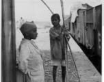 Two boys hang out at train station, one with stalks of sugarcane in hand.  Scenes in India witnessed by American GIs during WWII. For many Americans of that era, with their limited experience traveling, the everyday sights and sounds overseas were new, intriguing, and photo worthy.